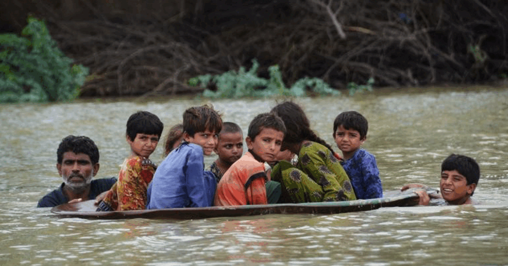 Children and a man traveling on a makeshift raft through floodwaters in a rural area, reflecting climate-related disaster impacts.